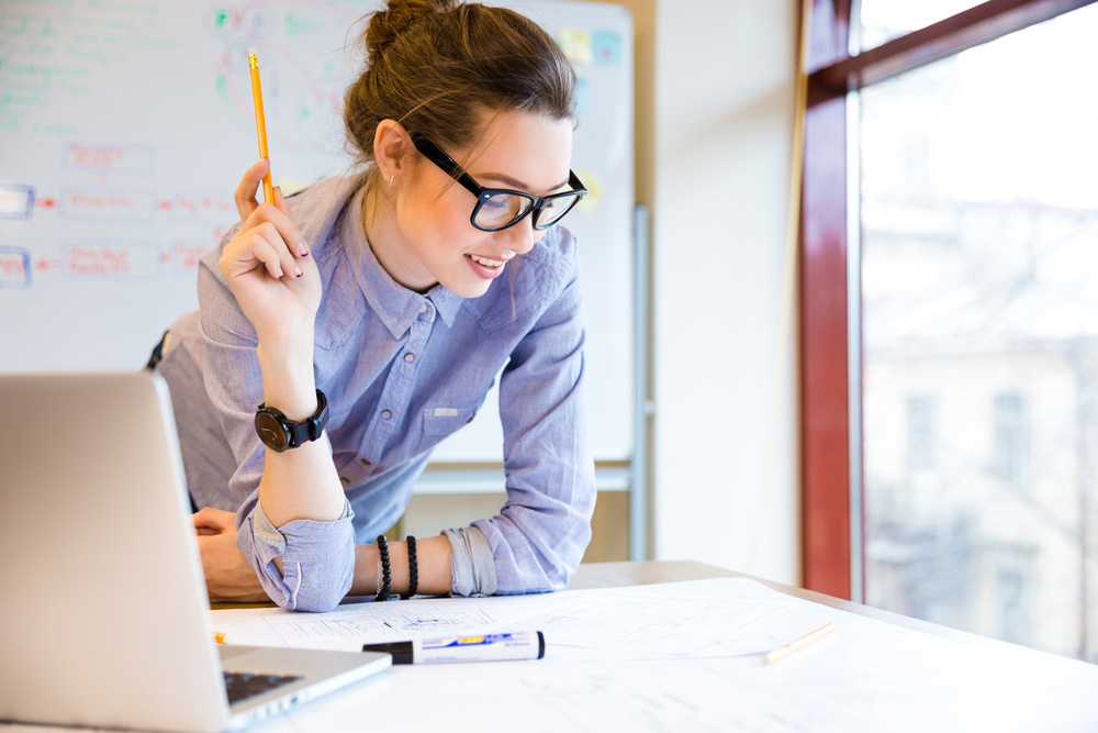 young woman in glasses standing near the window in office and working with blueprint